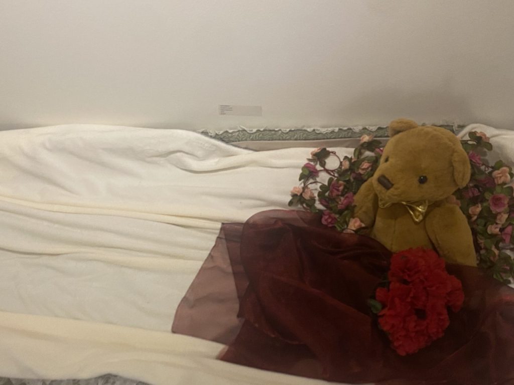 A medium-sized open coffin on the floor with a white blanket or spread laying across its bottom. It has a teddy bear and flowers with a red veil at the head of it.