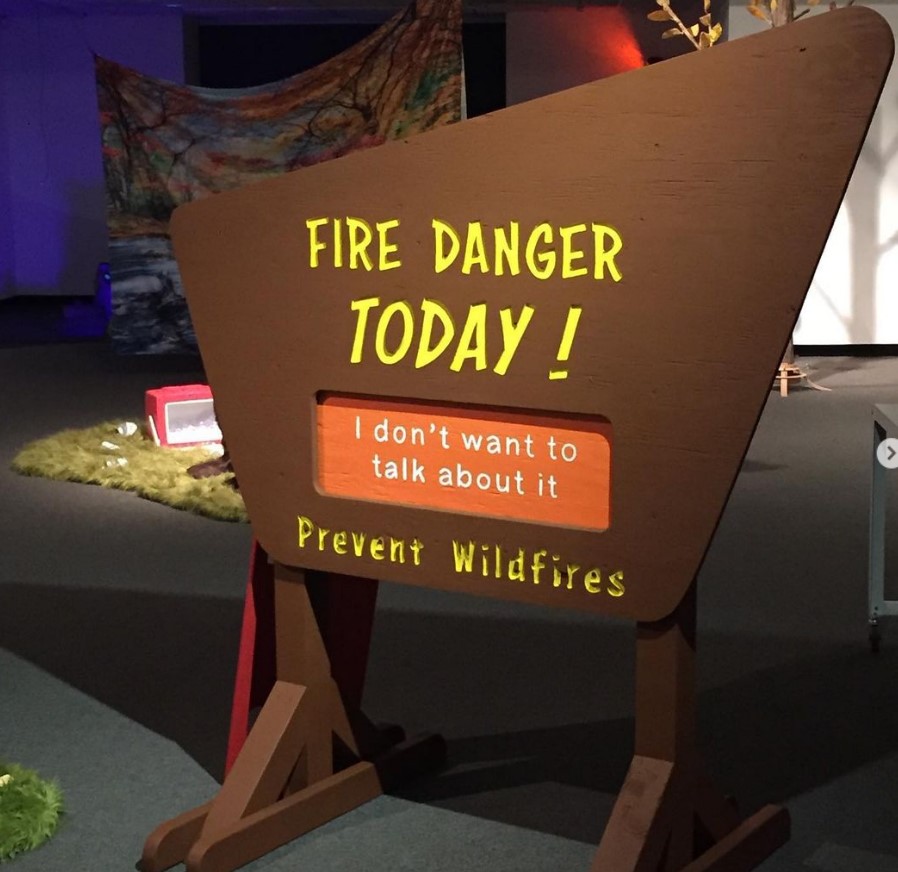 Wooden brown sign. Reads "fire danger TODAY!" "I don't want to talk about it" on an orange board that is incased in the brown sign. "Prevent wildfires" at the bottom of the brown sign.