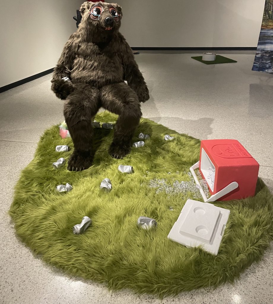 A sculpture of a fake bear sitting in a chair and holding empty cans. 