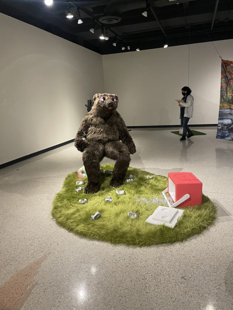 Drunken bear sitting on a lawn chair surrounded by drunken beer cans thathave fallen out of a tipped over cooler on a patch of wild grass.