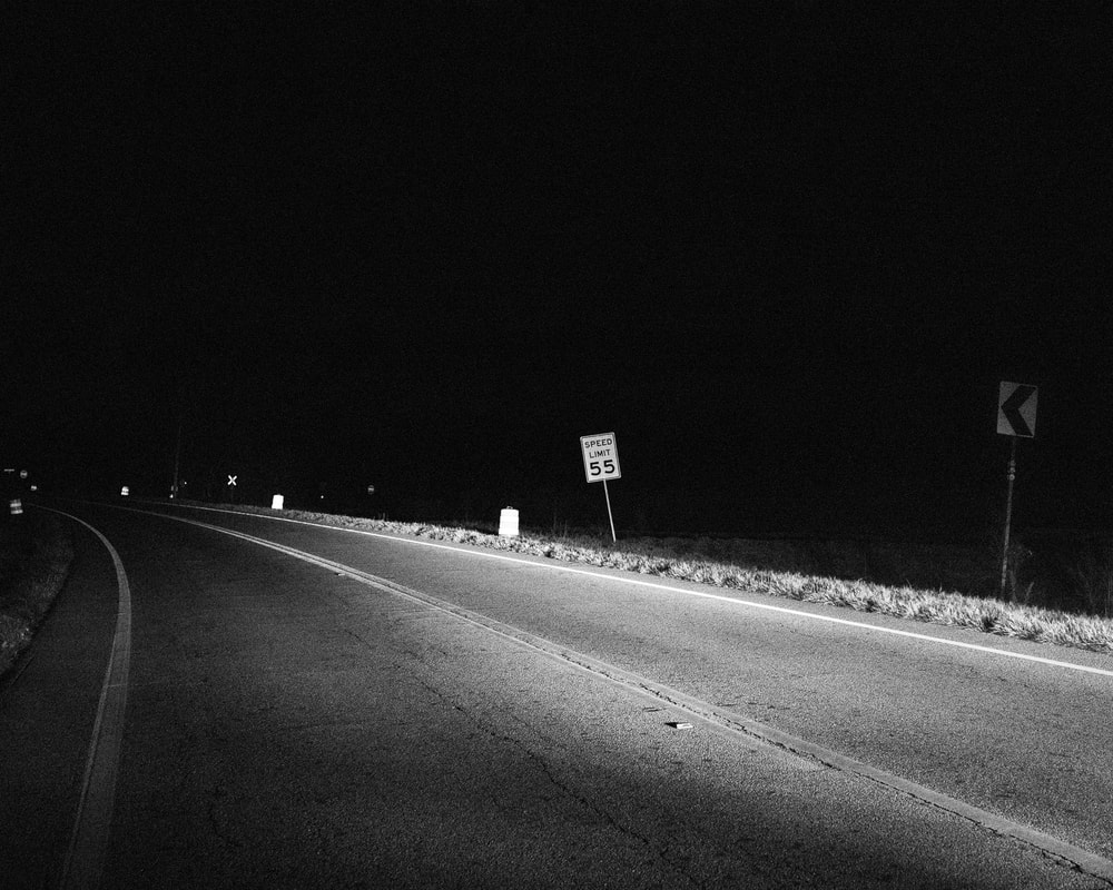 A photograph of a road taken at night with the right side of the road being illuminated by headlights. This includes, but is not limited to: A Speed Limit sign, a Train Track Crossing Sign, and an Arrow Sign.