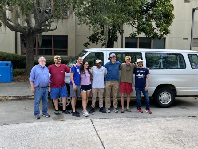 Students and Dr. Mark Groszos stand in front of a van outside of Nevins Hall on the campus of VState after returning from their trip.